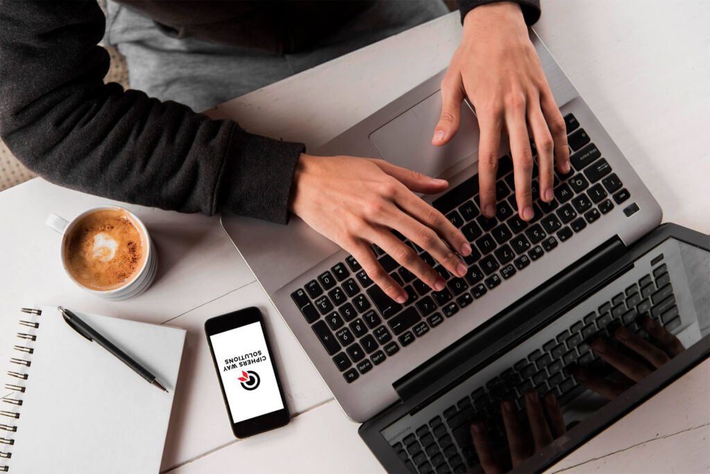 Close-up of a man's hands researching the market on a laptop, with a phone, coffee, notebook, and pen on the desk. The phone displays the Cipher Sway Solutions logo.