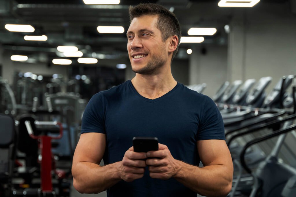 man in his early 30s walking through a gym, smiling while looking at his phone.